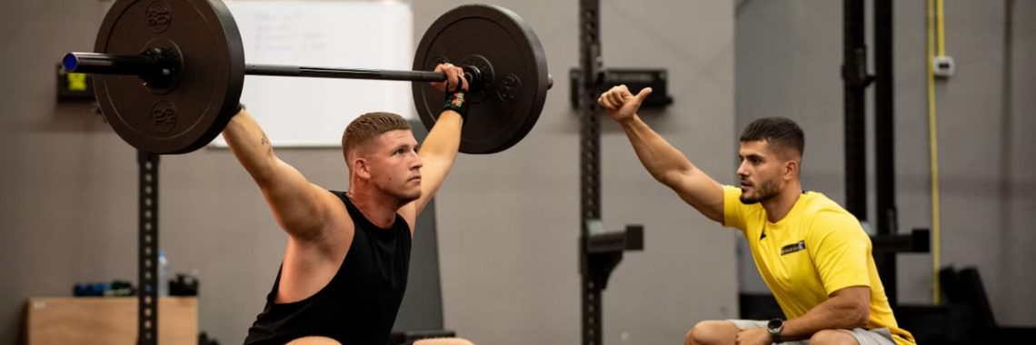 Man lifting weights under coach supervision in a gym setting, focusing on strength training.
